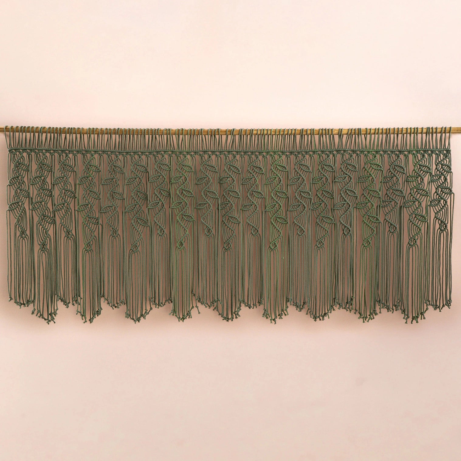 An olive green macrame wall hanging with a woven vine pattern and long fringe, suspended from a wooden rod against a plain beige wall.